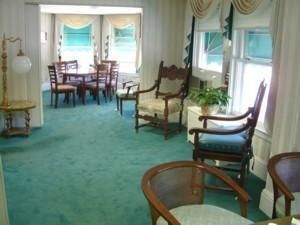 Bright living room with turquoise carpet, white curtains, and wooden chairs near large windows