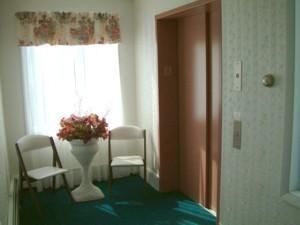 Small sitting area with two white chairs, a flower table, and an open elevator doorway in a hallway.