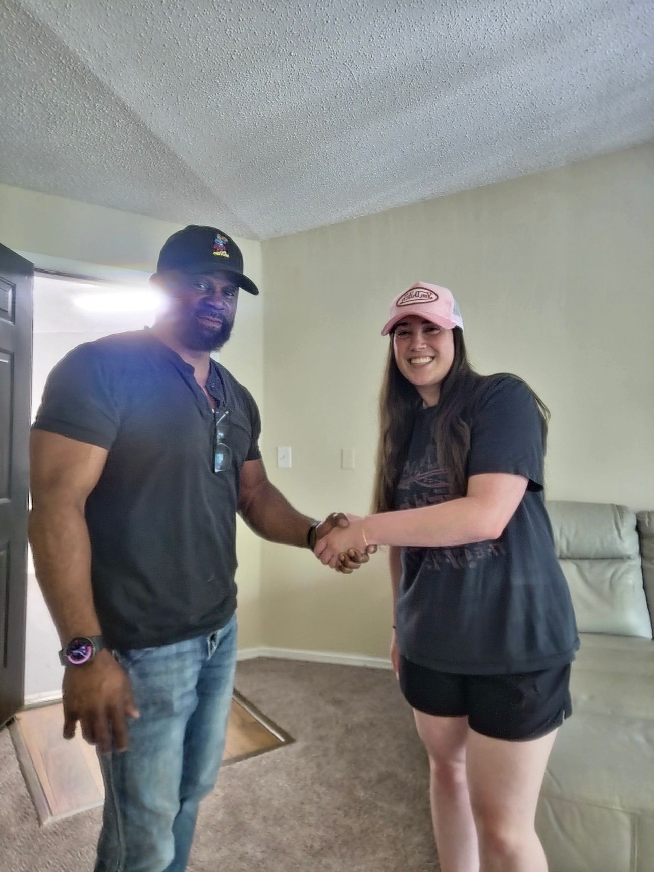 Two people shaking hands indoors. Man in black shirt, jeans, and cap. Woman in black shirt, shorts, and pink cap.