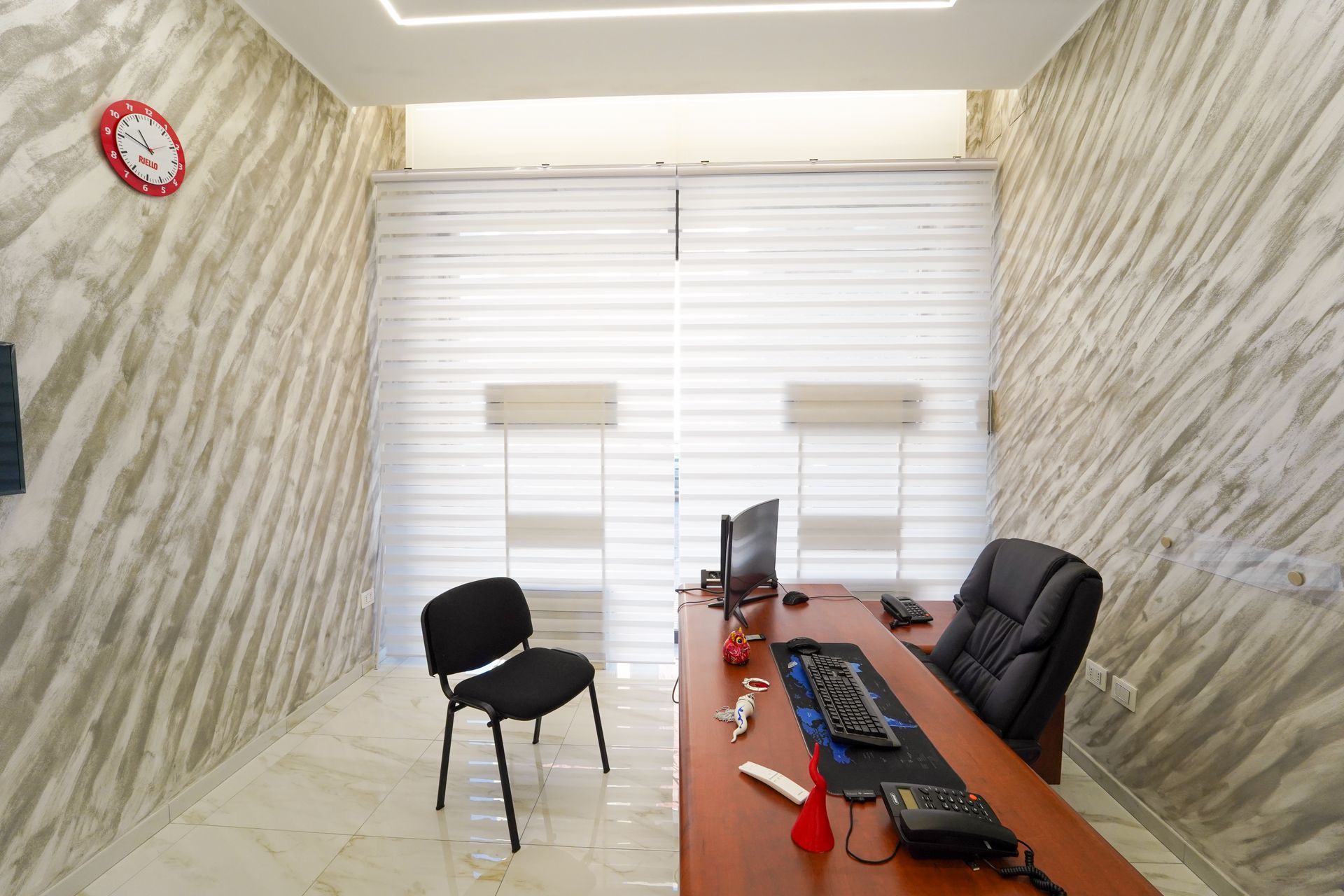 Interno di un ufficio con scrivania, sedia, computer, tende, pareti strutturate e un orologio.