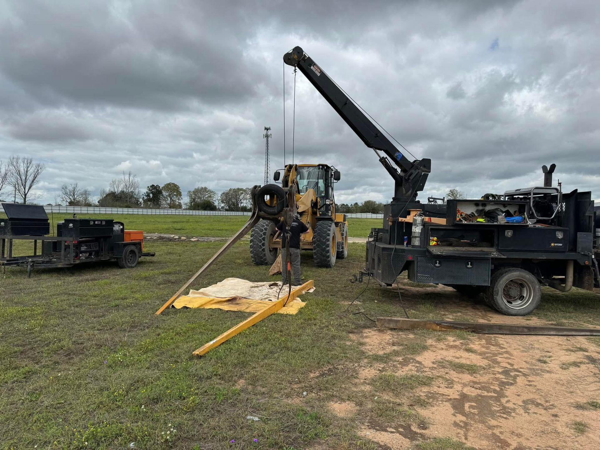 A truck with a crane, a yellow bulldozer, and a trailer on a green field under a cloudy sky. Precision Parts Manufacturing