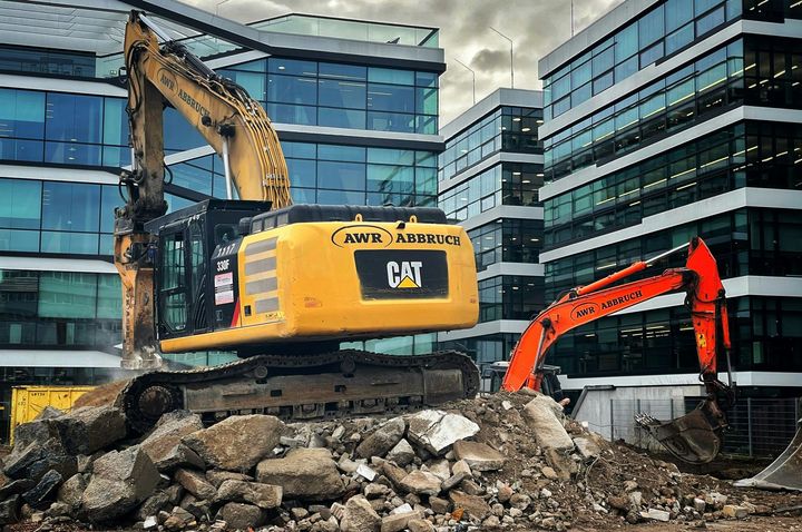 A yellow CAT excavator and an orange excavator working on a pile of rubble in front of modern glass office buildings.
