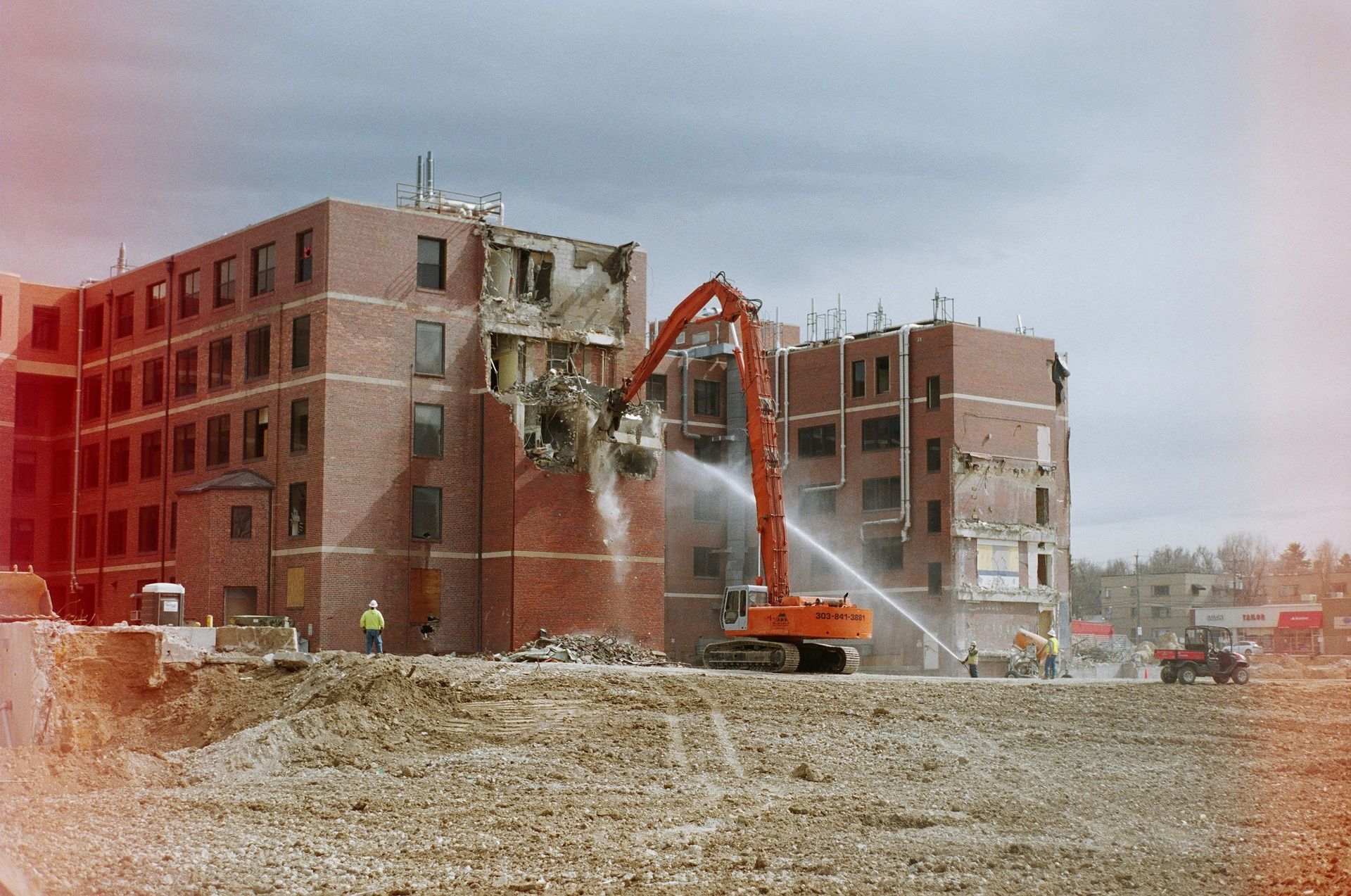 An orange excavator demolishes a multi-story brick building while spraying water to control dust on a construction site.