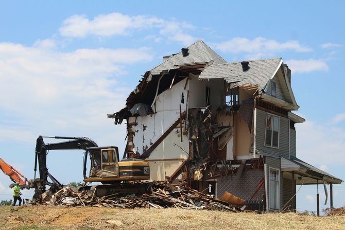 A large excavator demolishes a house, leaving a pile of debris against a clear blue sky.