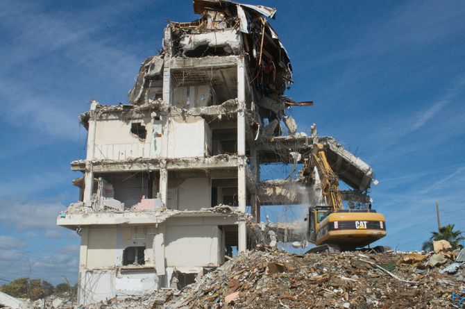 A yellow excavator tears down a multi-story concrete building amid a pile of rubble under a clear blue sky.