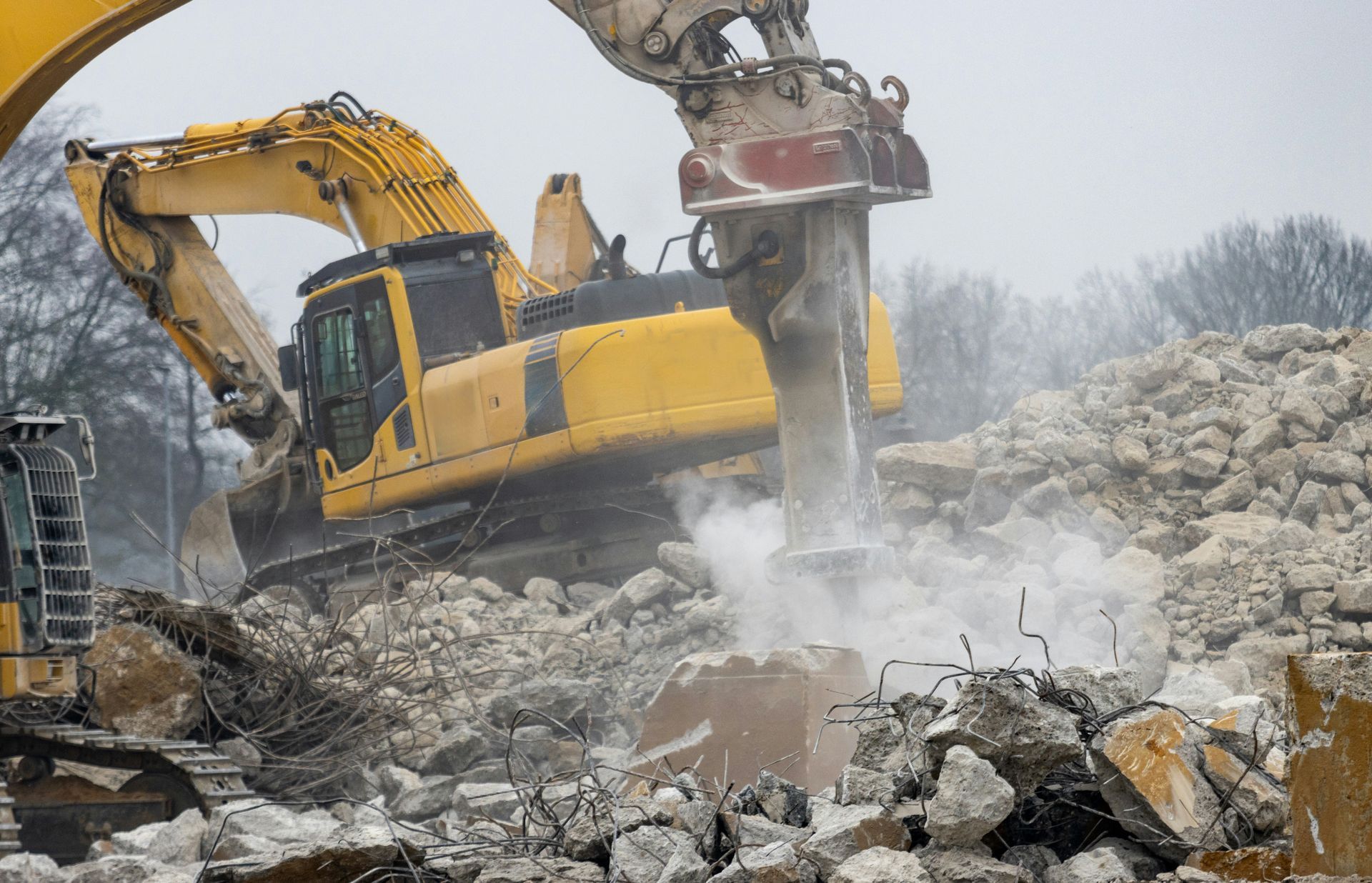 A yellow excavator equipped with a hydraulic hammer breaks through a pile of concrete rubble at a demolition site.