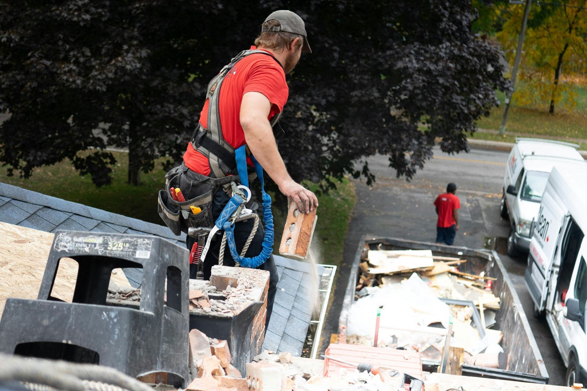A person in a red shirt and tool belt tosses a brick into a disposal bin from a rooftop construction site.