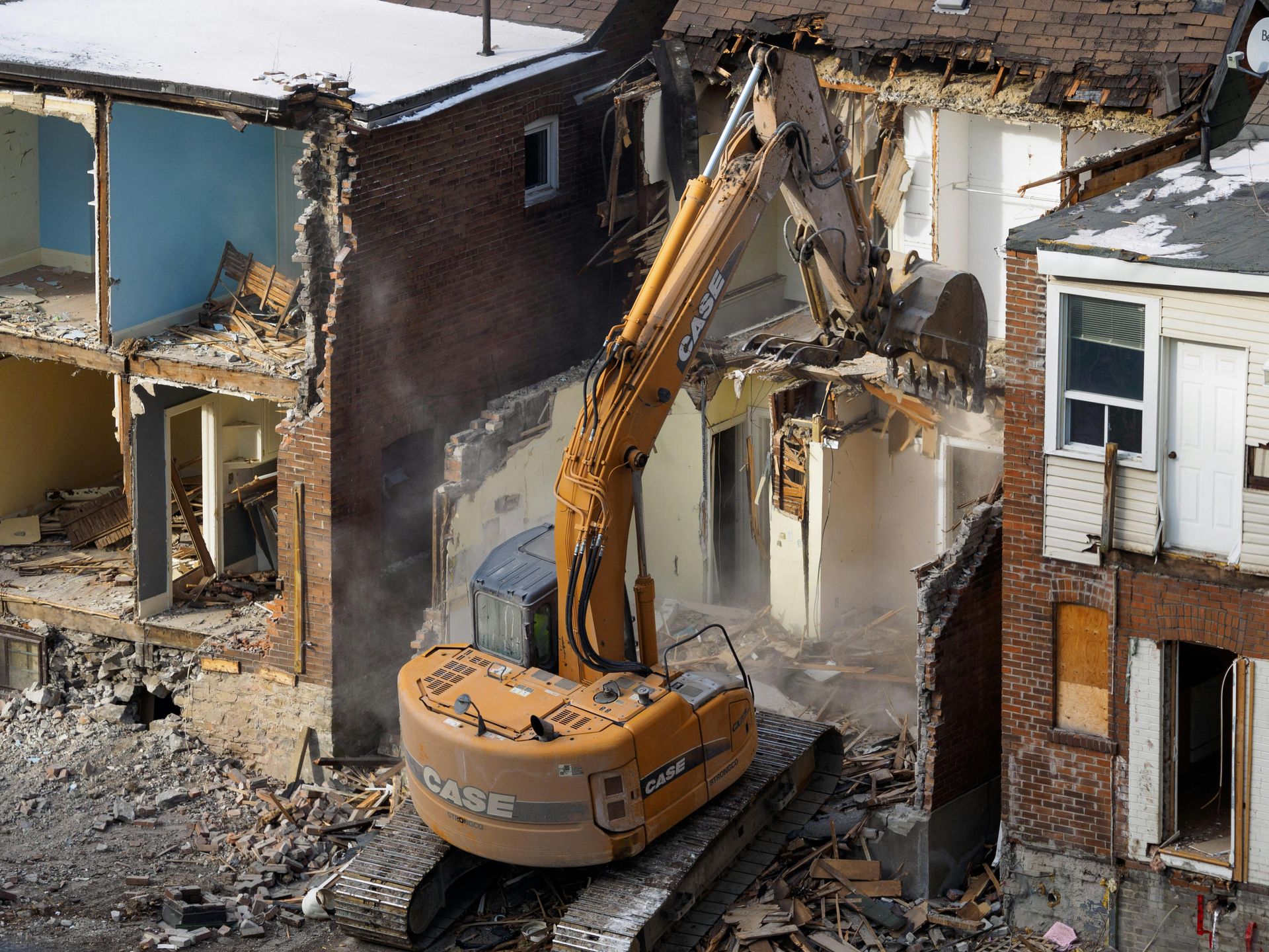 A yellow CASE excavator demolishes a brick residential building, exposing interior walls and debris.