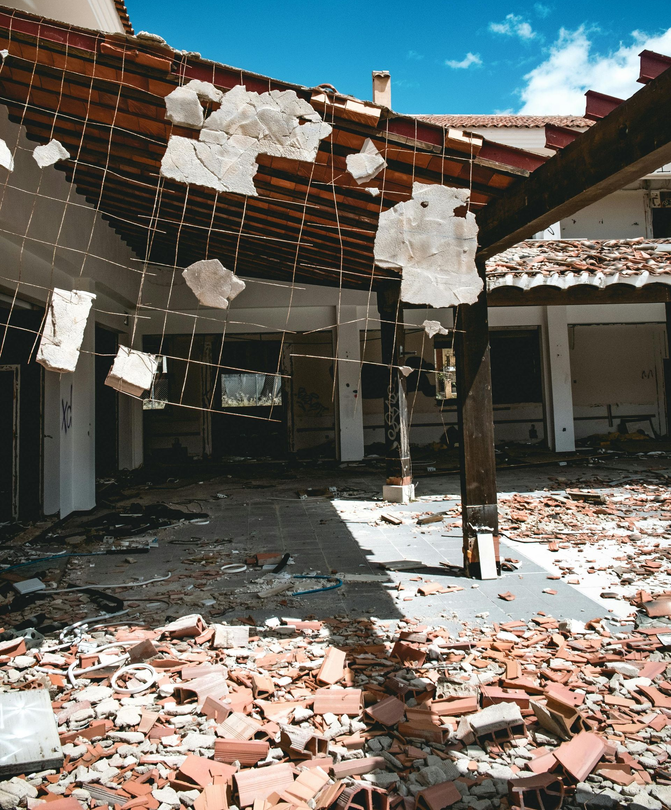 A partially destroyed building interior with a damaged roof, exposed wooden beams, and rubble covering the floor.