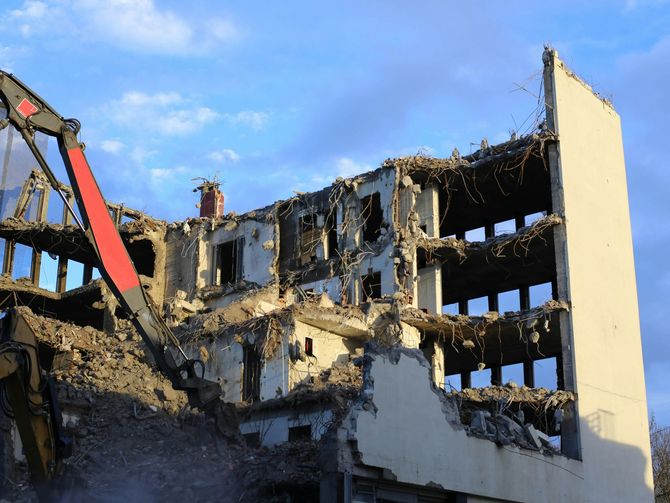 A large construction excavator demolishing a multi-story concrete building under a blue sky.