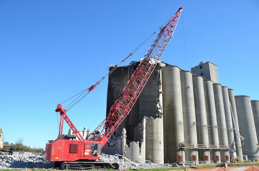 A large red crane with an extended boom stands beside a concrete grain silo that is partially demolished.