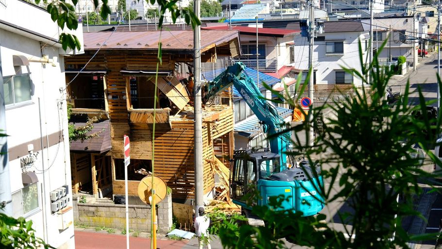 A teal excavator dismantles a wooden structure near a residential street on a sunny day.