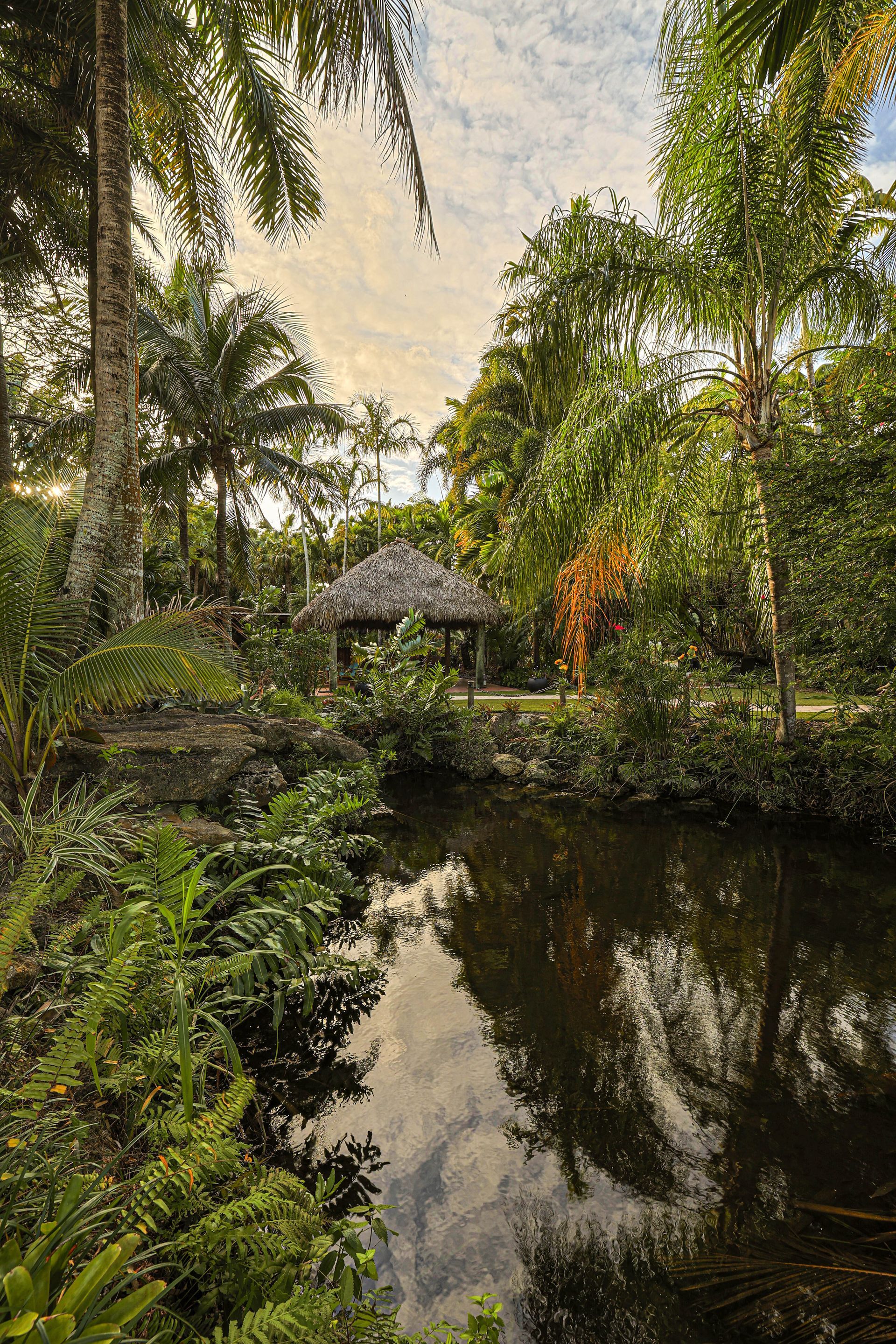 A serene pond reflecting lush green trees and foliage, with a small, thatched-roof gazebo nestled in the background.