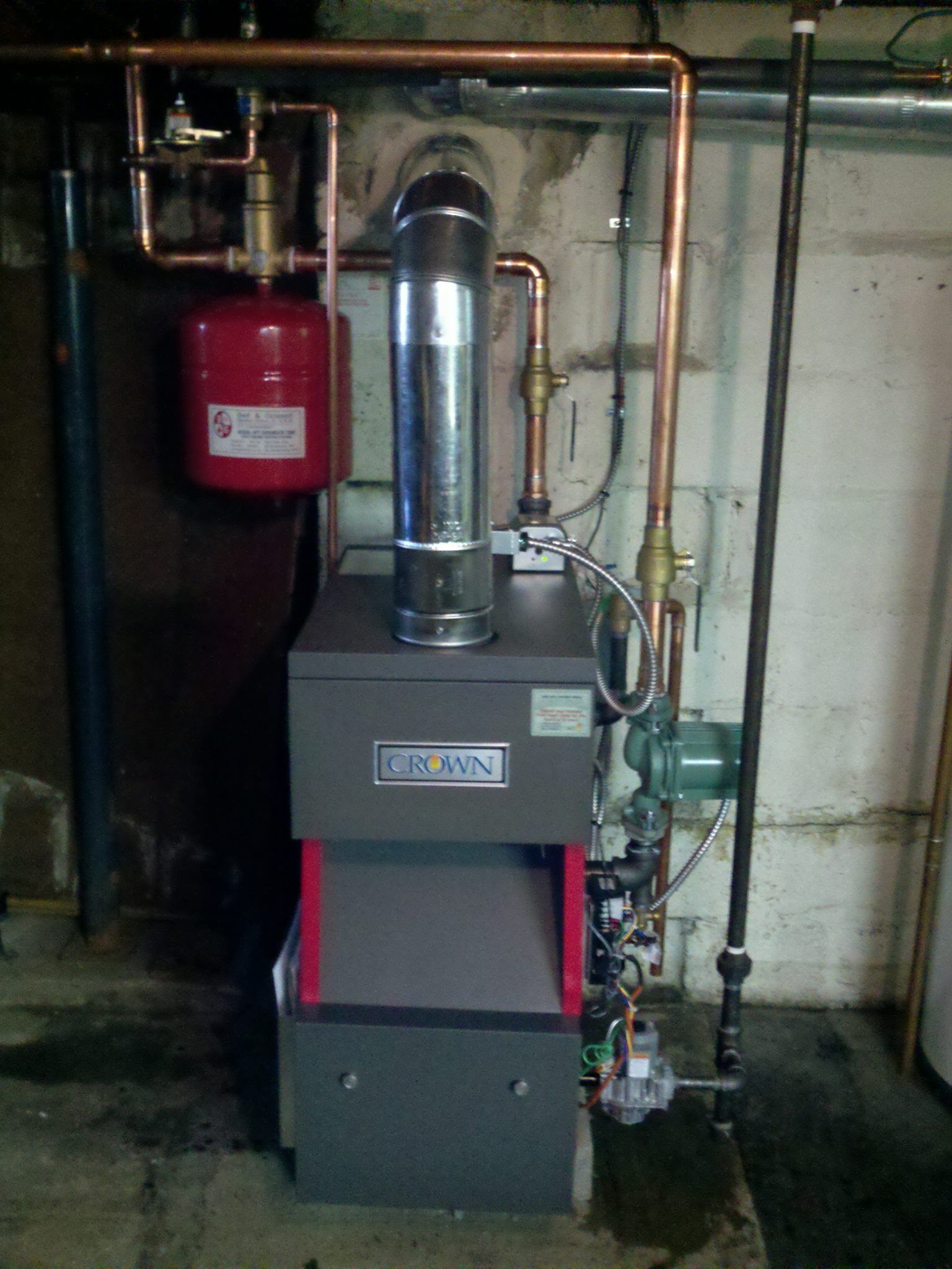 A gas-fired boiler with red expansion tank, copper pipes, and metal chimney in a basement setting.