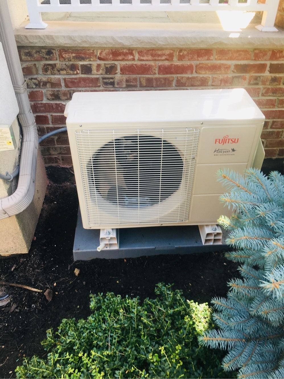 Fujitsu air conditioning unit on a concrete base, near brick wall and blue shrubbery.