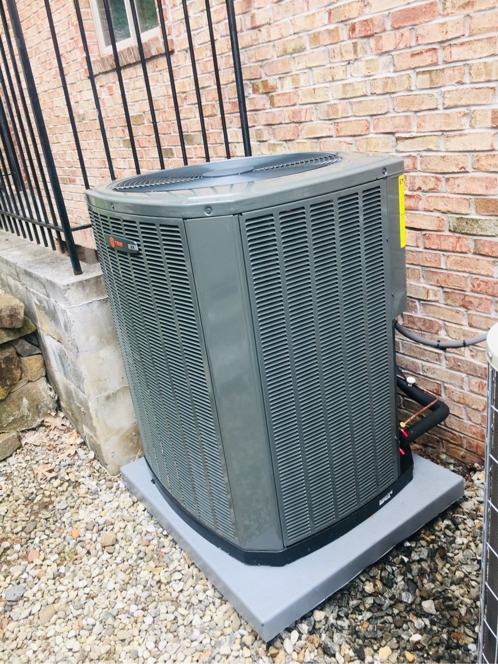 Gray Trane air conditioner unit next to a brick wall and a black metal fence on a concrete pad.