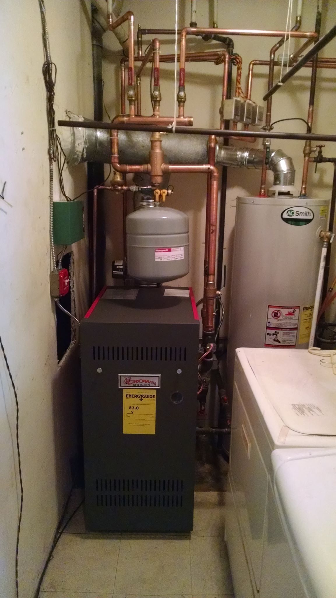 Furnace, water heater, and piping in a basement utility room; copper pipes, gray appliances, white wall.