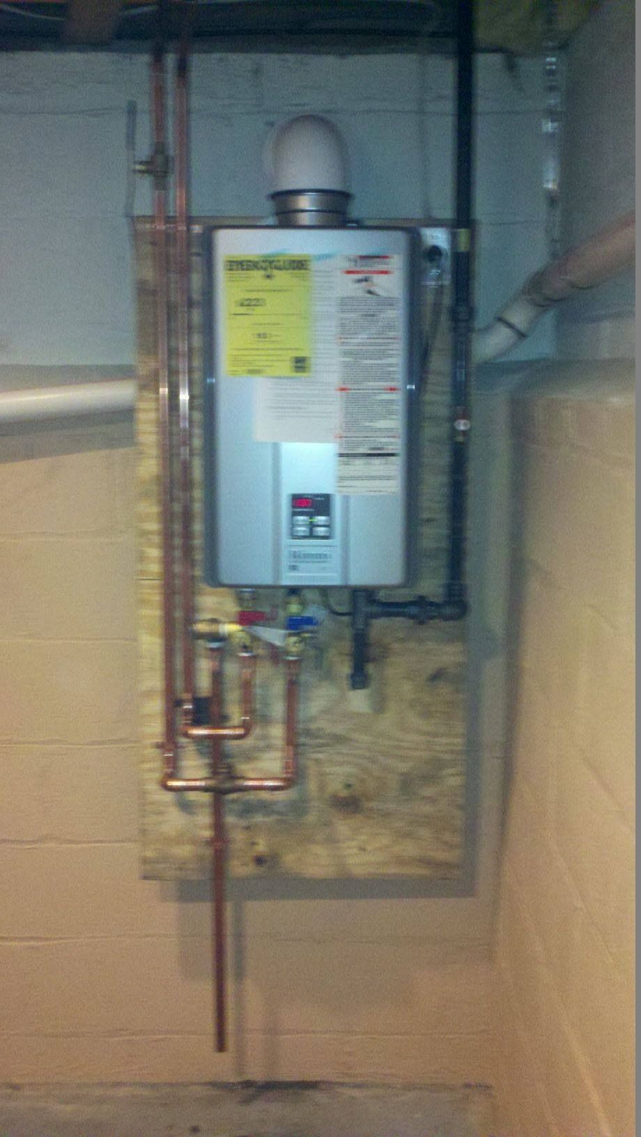 Tankless water heater mounted on plywood in a basement, with copper pipes and a white vent.