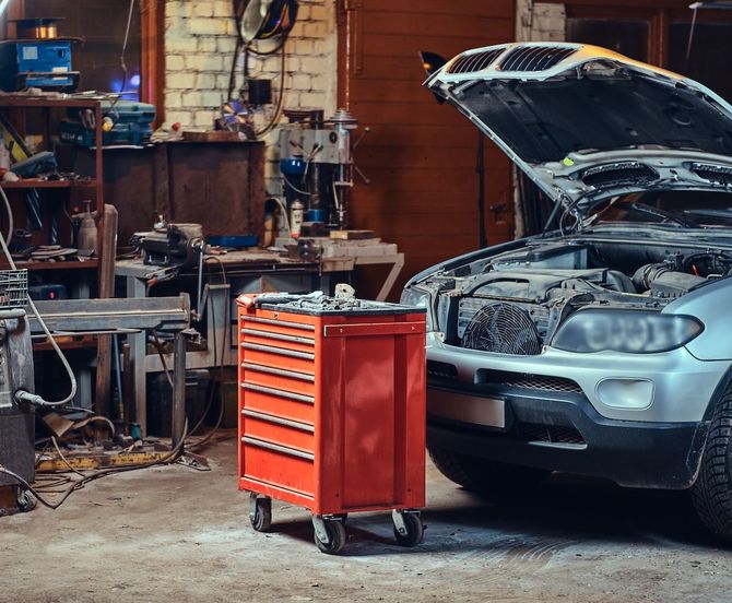 Orange tool chest in a cluttered auto garage beside a silver car with its hood open and engine exposed