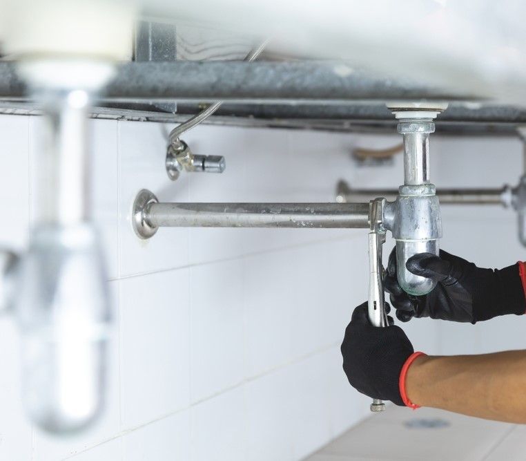 Plumber With Black Gloves Using Wrench To Repair Sink Pipes Under White Tiled Wall — David Talbot Plumbing in Cranbrook, QLD