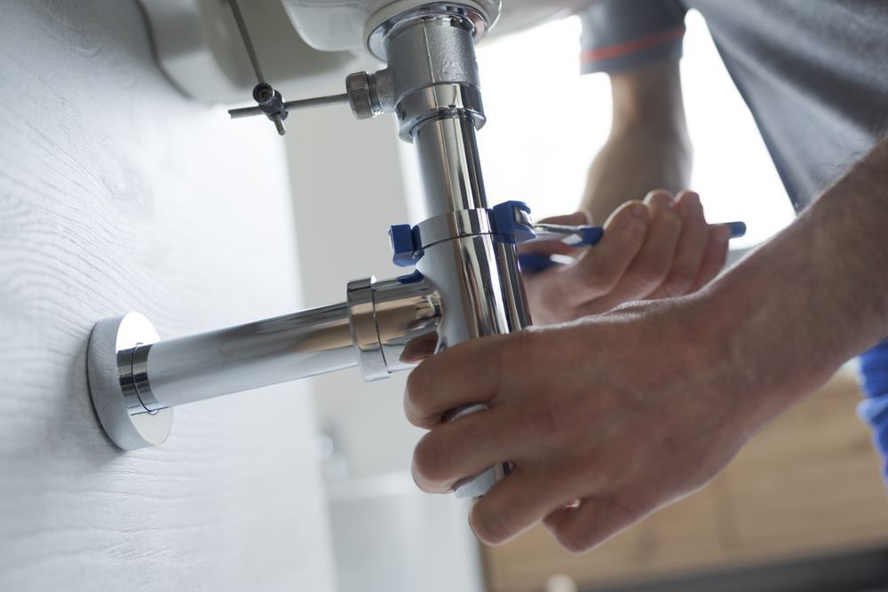 Plumber Using a Wrench to Tighten a Chrome Pipe Under a Sink in A Bathroom — David Talbot Plumbing in Cranbrook, QLD