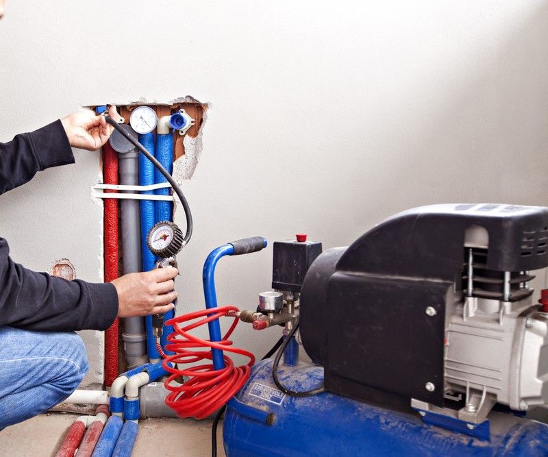 Plumber Checking Pipes With Gauge Near Blue Air Compressor Indoors — David Talbot Plumbing in Cranbrook, QLD