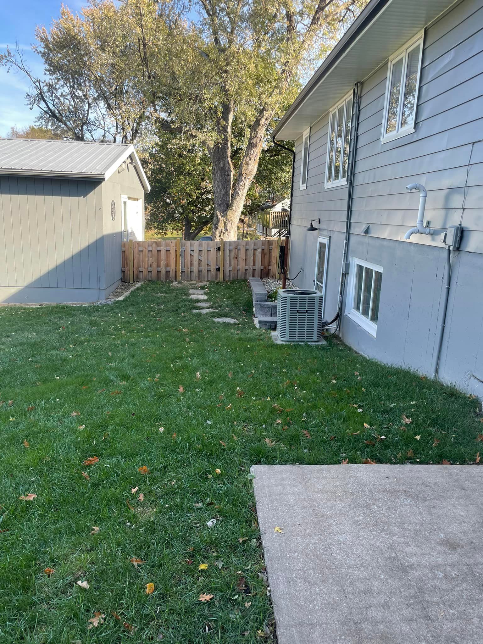 Backyard lawn beside a gray house, with a shed, air conditioner, fence, and large tree.