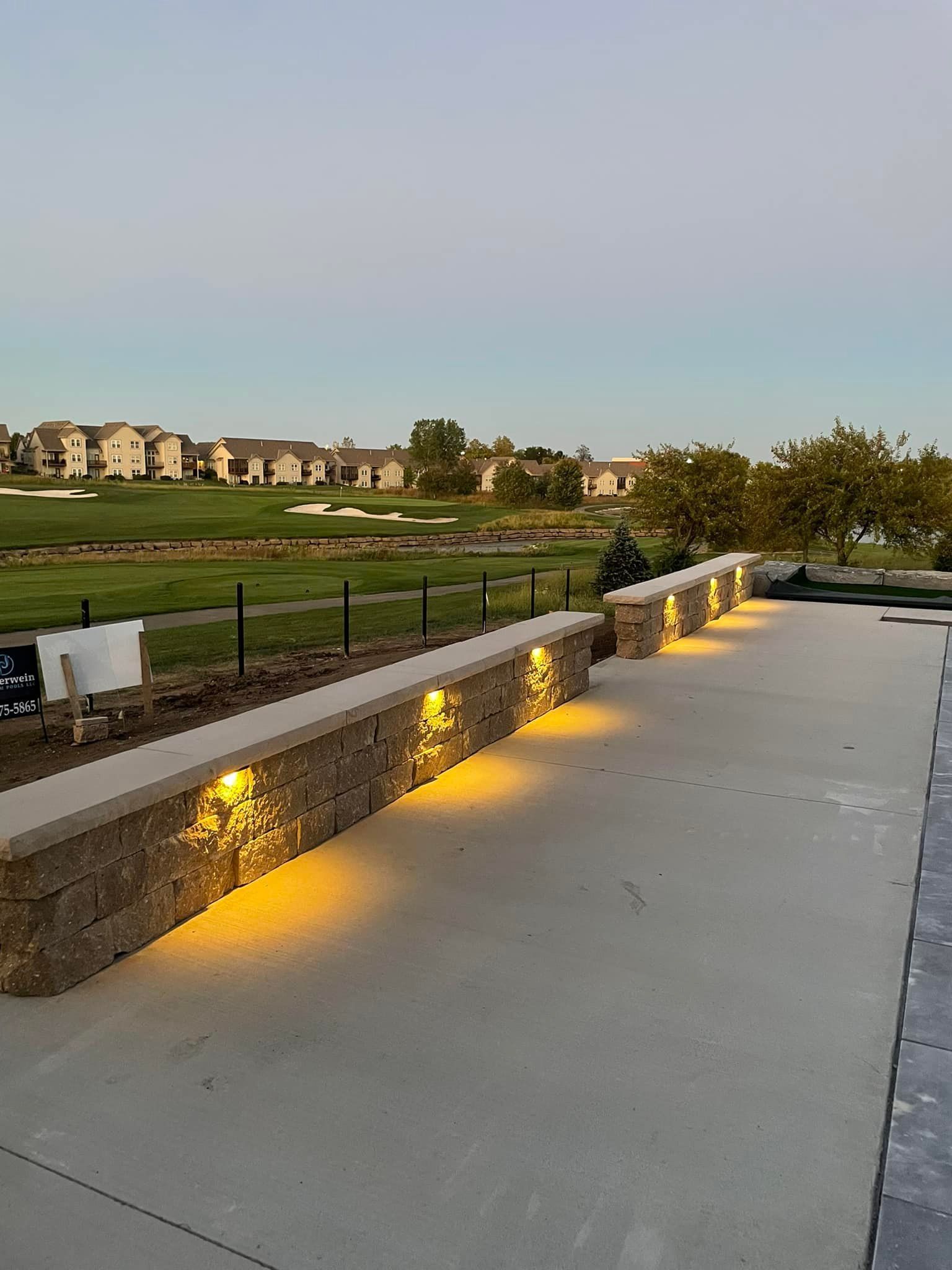 Stone walkway with warm ground lights beside a grassy park at dusk, buildings in the distance