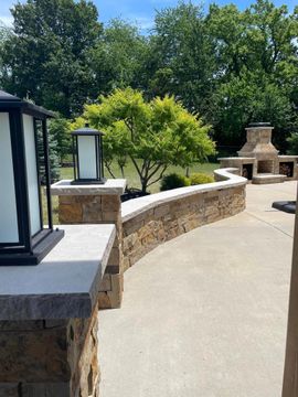 Stone patio walkway beside a low wall and outdoor fireplace, with green trees in the background.