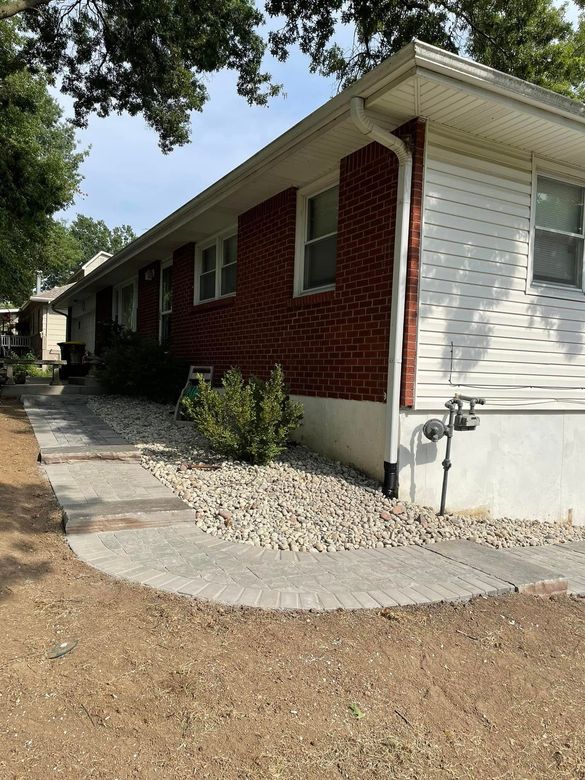 Single-story brick house with white siding, side walkway, and gravel landscaping under trees