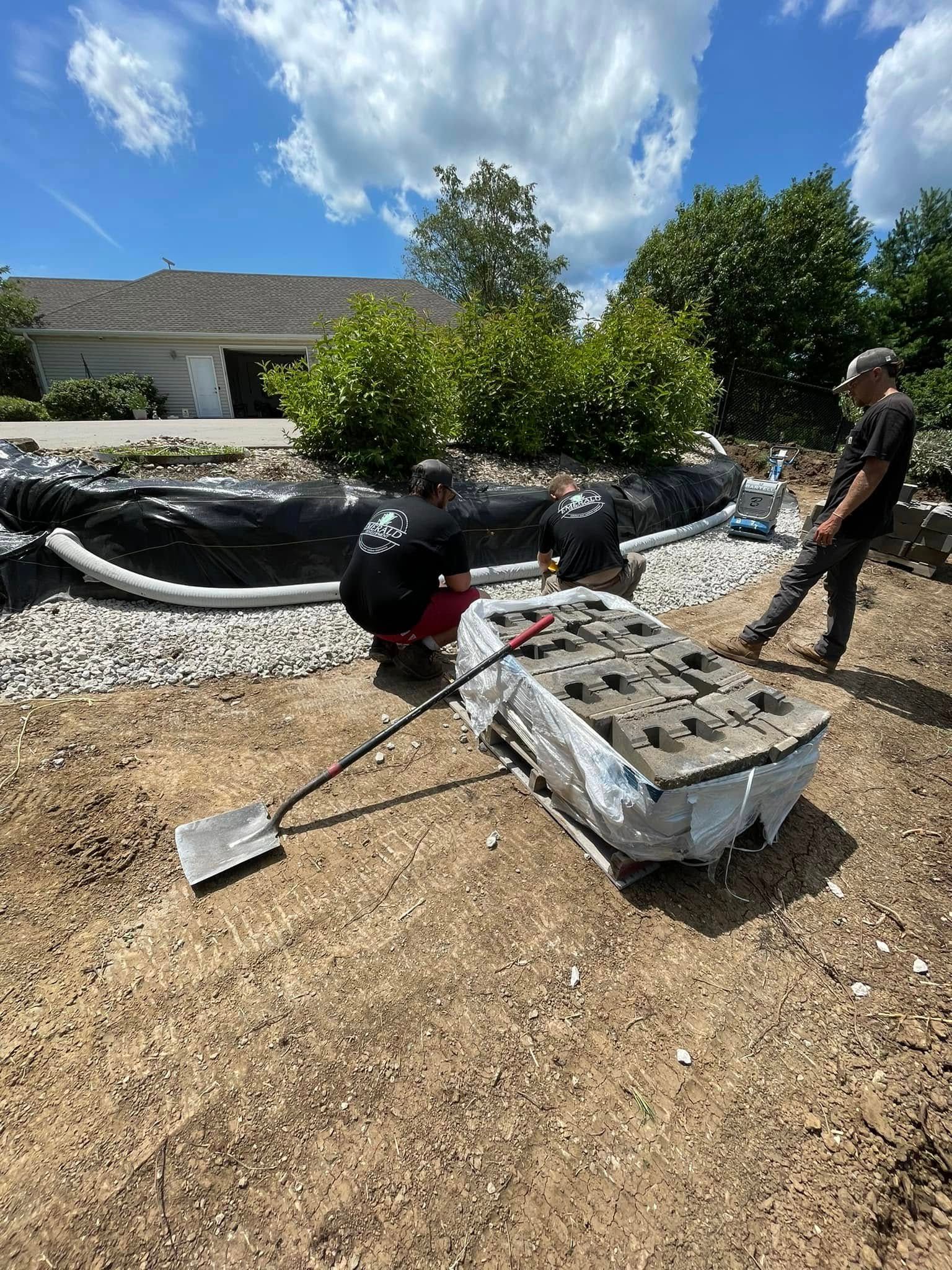 Workers tend a black sculpture beside a white stone bed in a sunny outdoor garden area.