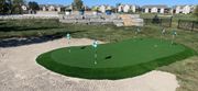 Small artificial putting green with golf flags beside a gravel path in a suburban neighborhood.