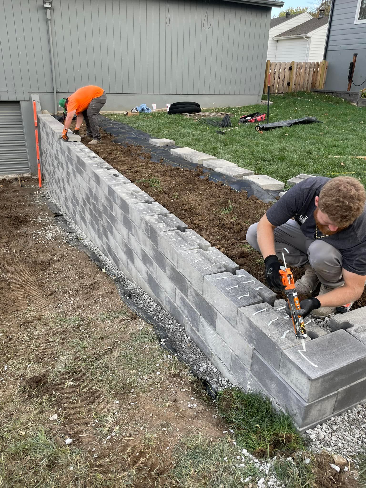 Two workers building a gray retaining wall along a grassy yard with a house in the background