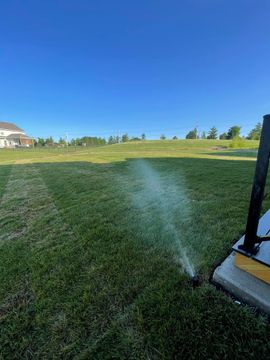 Sprinkler spraying water over a green lawn beside a black fence under a clear blue sky