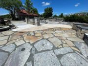 Stone patio with low walls and a pergola under a bright blue sky, surrounded by trees and landscaping