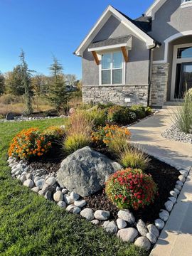 Landscaped front yard with colorful flowers, rocks, and a stone walkway beside a gray house