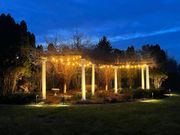 Lit pergola in a dark garden, glowing with warm string lights and surrounded by trees.