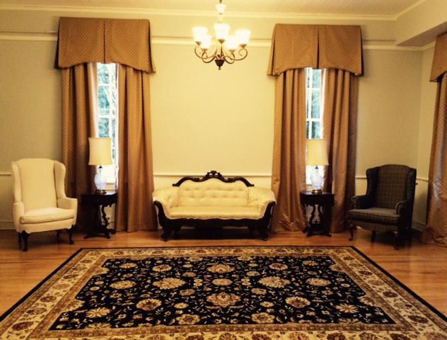 Elegant living room with ornate rug, cream and dark chairs, sofa, and tall curtains under a chandelier