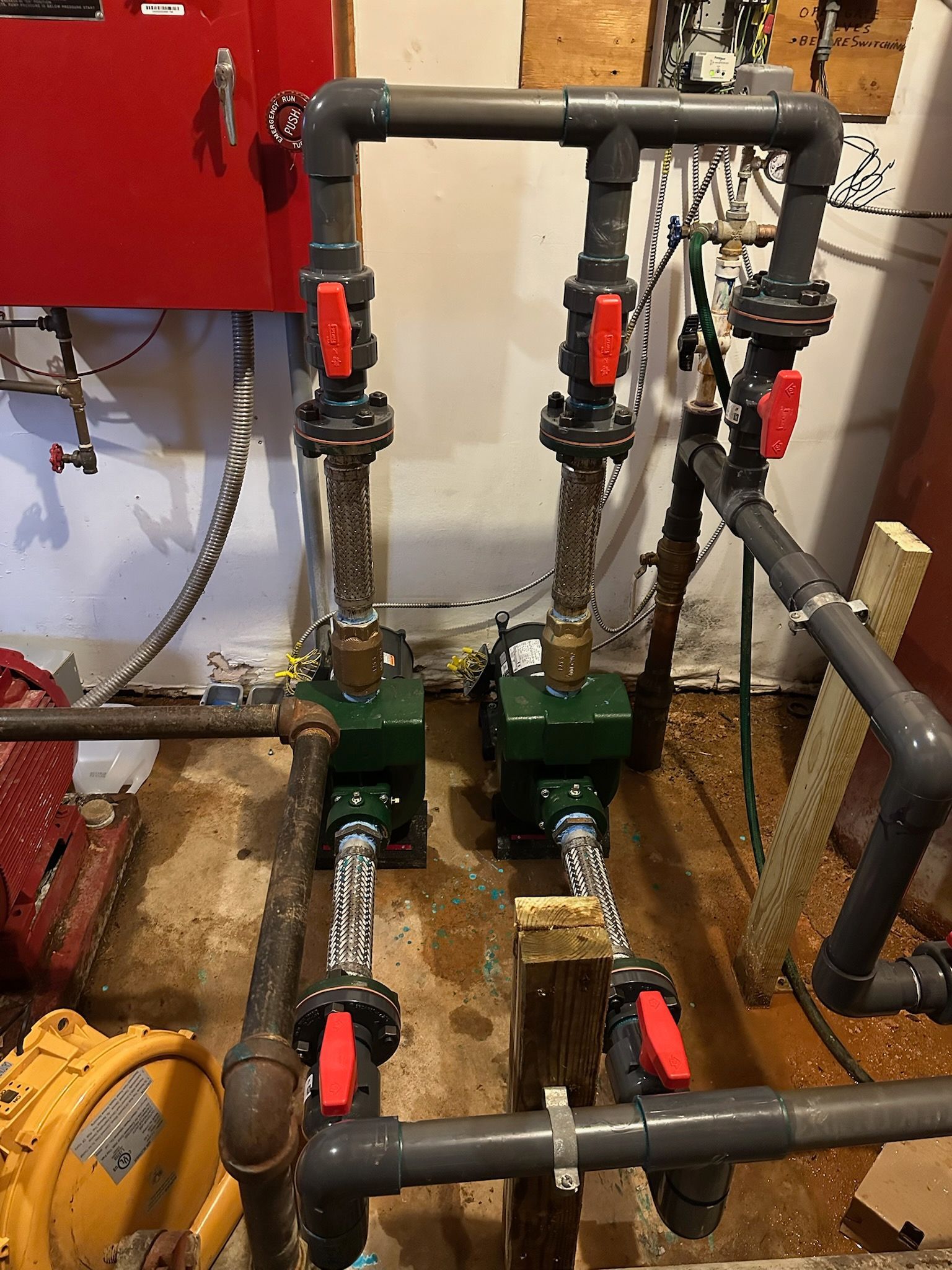 Piping system with two green pumps, red valves, and grey pipes in a utility room.