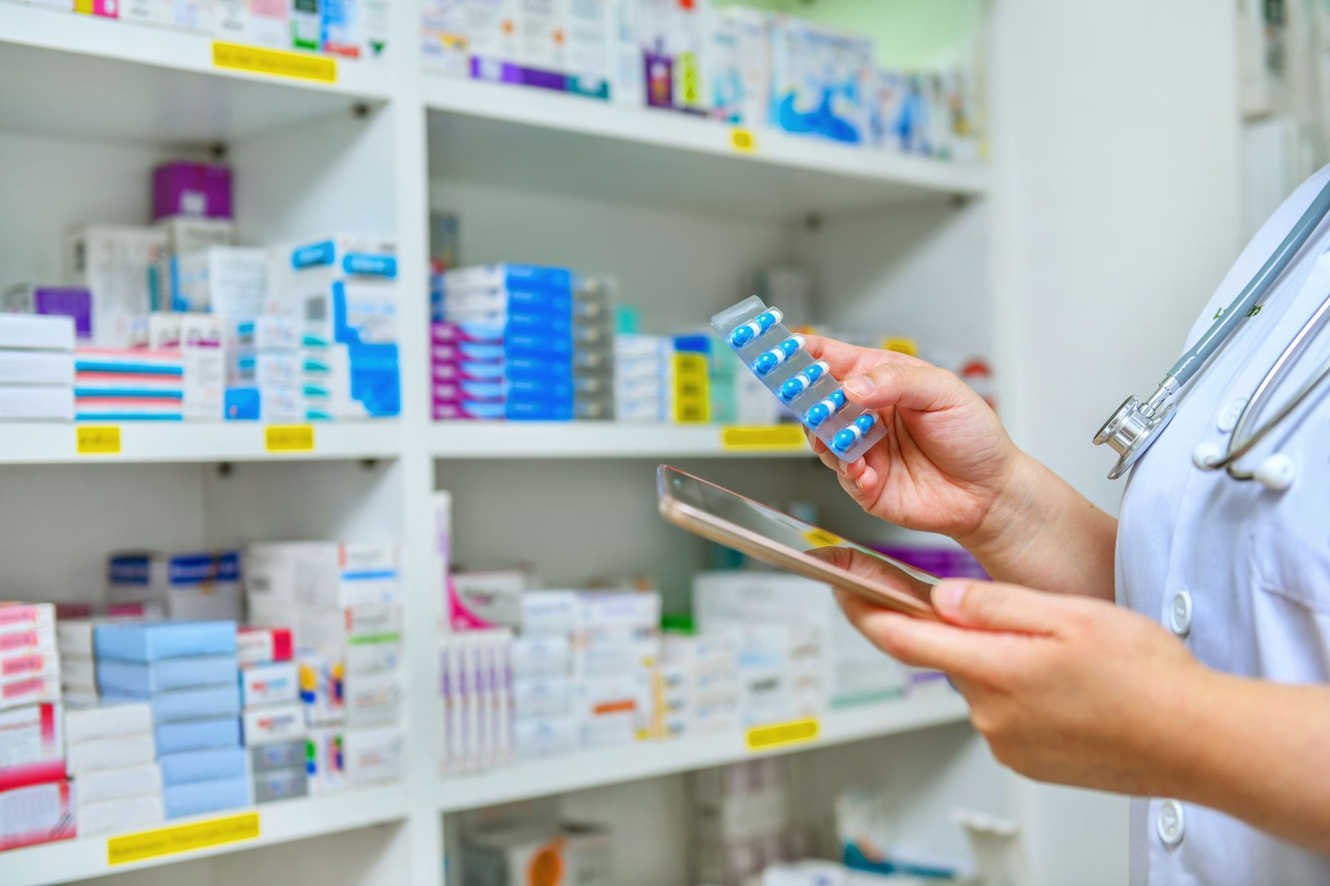 Pharmacist holding medication blister pack, consulting a tablet, in a pharmacy setting with stocked shelves.