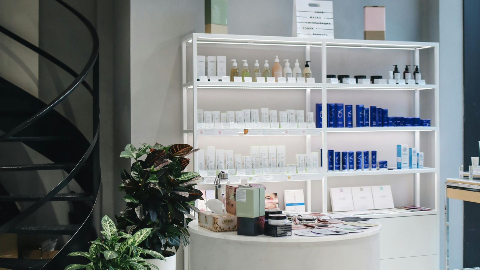 A display of skincare products on white shelves in a retail space with a spiral staircase and plants.