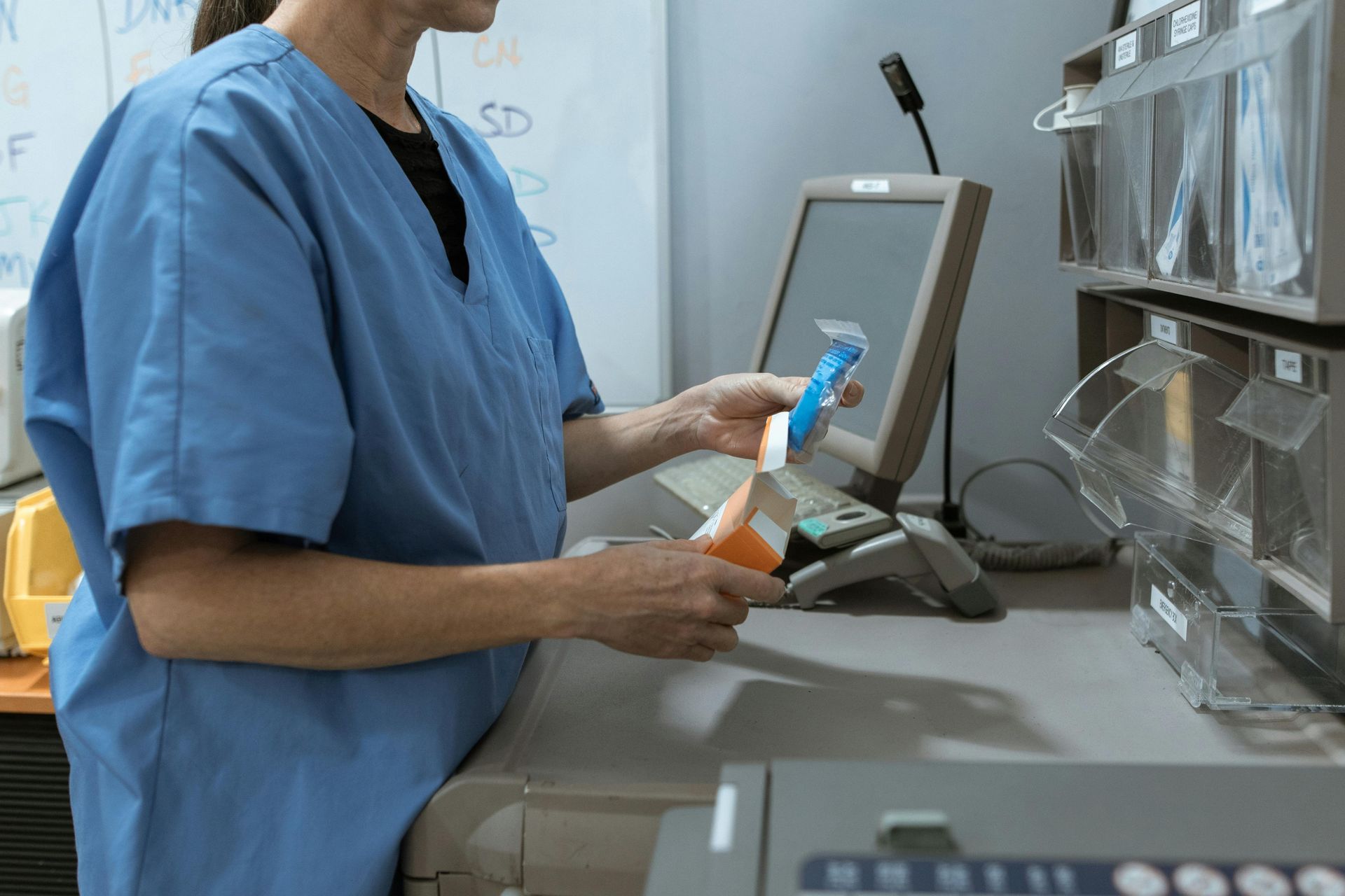 Pharmacy worker in blue scrubs holding medication bottles at a counter with a computer and storage bins.