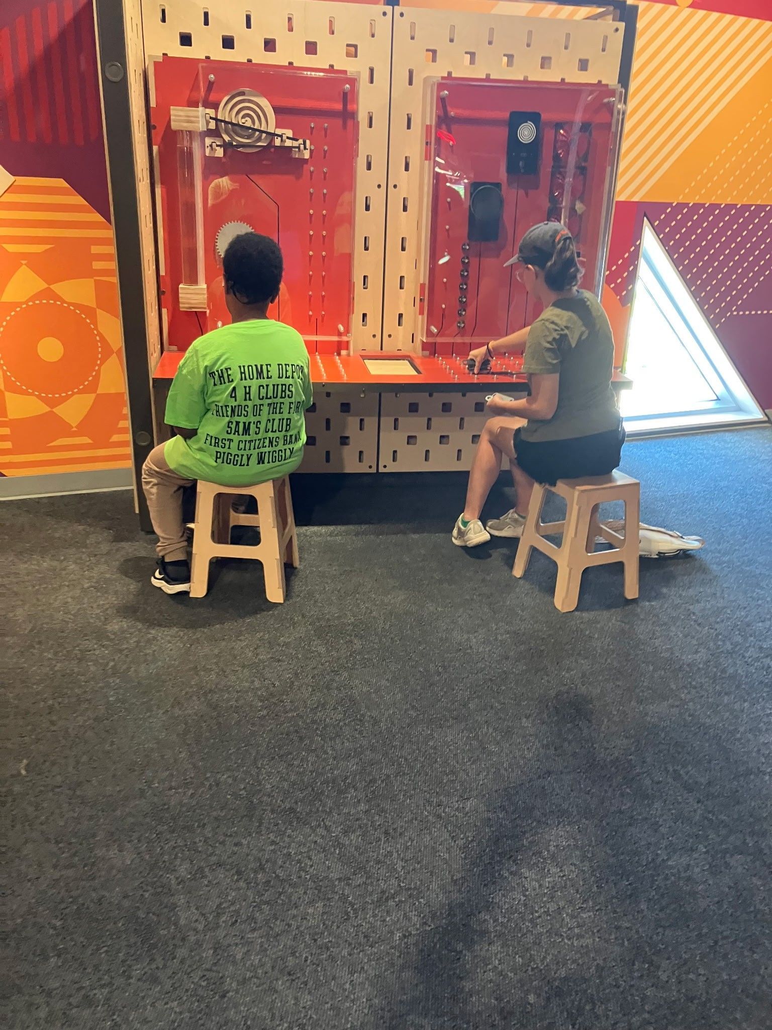 Two people sit on wooden stools in front of a red, interactive wall display in a brightly patterned room.