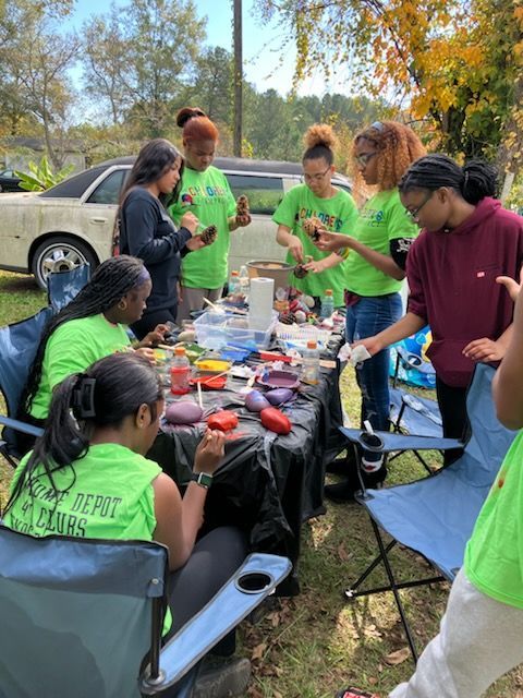 A group of people gathers outdoors around a table covered in craft supplies to paint rocks on a sunny day.