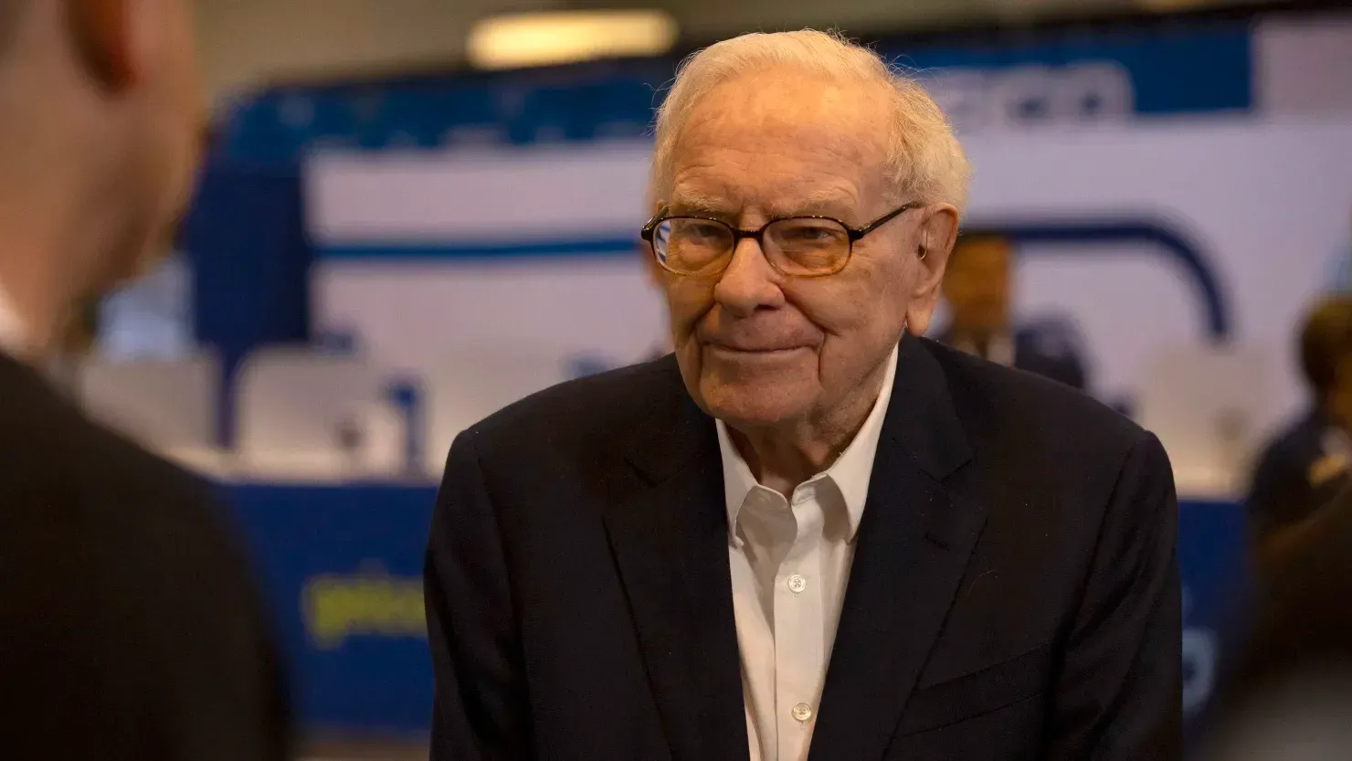 An older man wearing glasses and a suit is talking to another man.