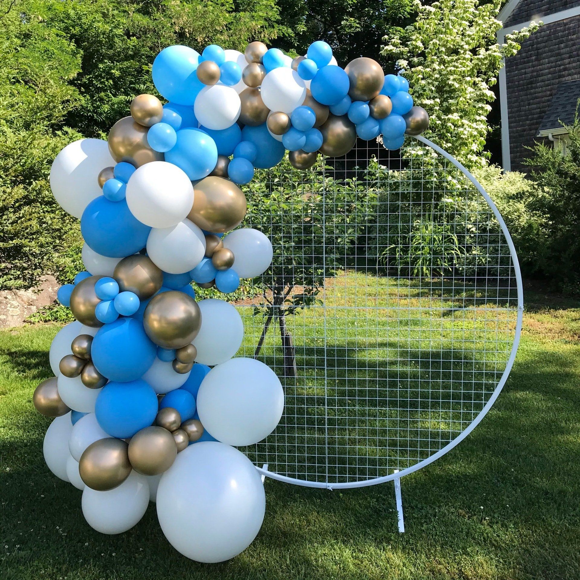 Pale blue, white, and  reflex gold  Organic Balloon garland on a white circular grid backdrop wall set on grass.