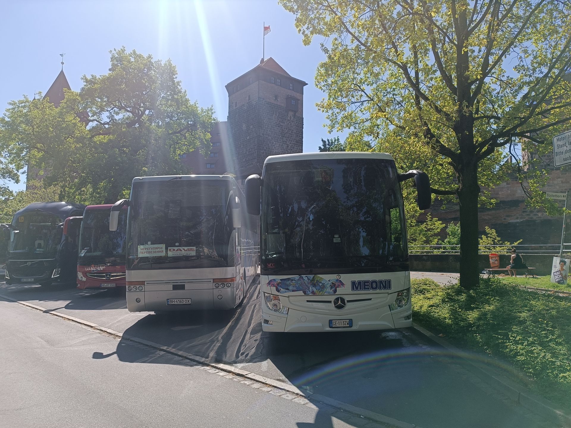 Tre autobus bianchi parcheggiati lungo una strada soleggiata, accanto ad alberi e una torre di pietra.