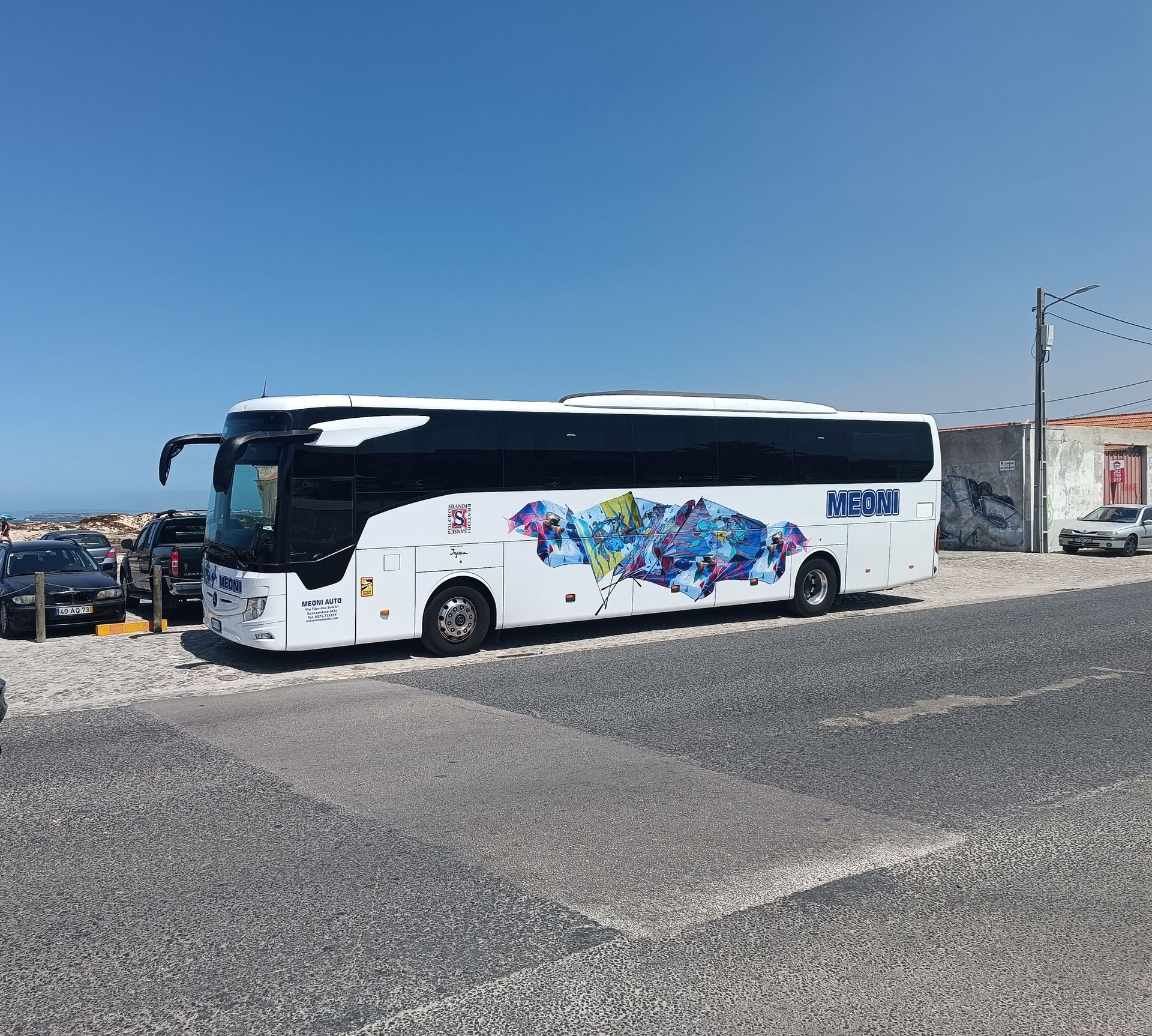 Autobus bianco con grafiche laterali colorate parcheggiato su un piazzale ghiaioso sotto un cielo azzurro e limpido.