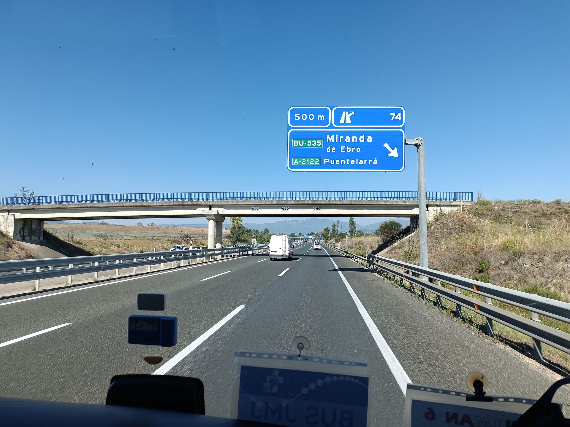 Vista dell'autostrada sotto il ponte segnalato con un cartello blu, con un furgone bianco davanti e l'erba secca a bordo strada sotto un cielo limpido.