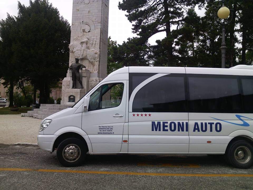 Furgone bianco MEONI AUTO parcheggiato vicino a un monumento e ad alcuni alberi in una piazza.